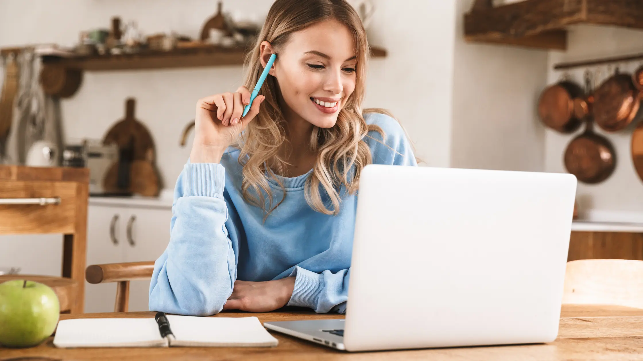 woman sitting at table with laptop and pen in hand, working while smiling