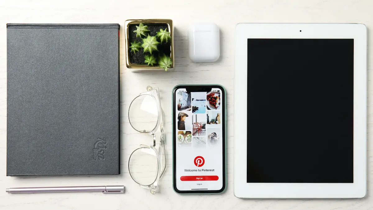 ipad, phone displaying pinterest, glasses, plant, and notebook laying on a table