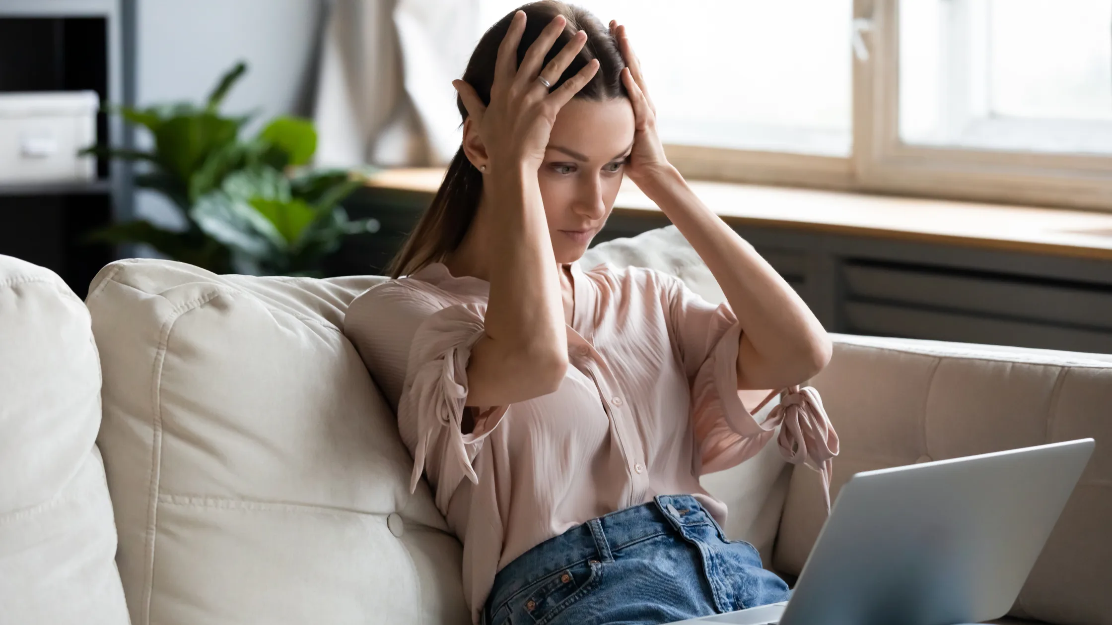 woman looking at computer, sitting on the couch. She is frustrated and is holder her hands on her forehead.